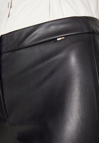 Gros plan sur la ceinture d'une jupe en cuir noire avec un petit détail rayé noir, blanc et beige, portée avec une chemise beige boutonnée.
