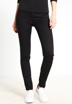 Femme portant un pantalon noir ajusté et des baskets blanches, debout avec les mains détendues sur les côtés devant un fond blanc uni.
