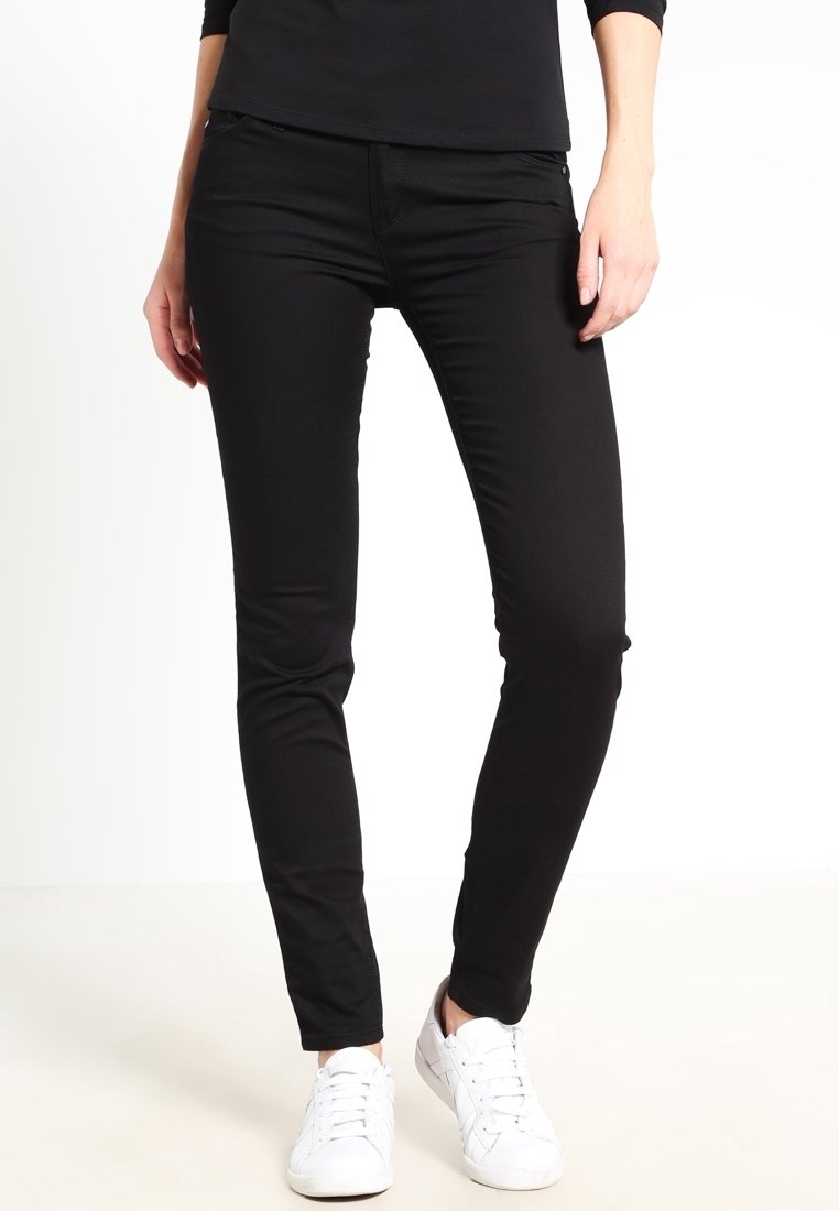 Femme portant un pantalon noir ajusté et des baskets blanches, debout avec les mains détendues sur les côtés devant un fond blanc uni.