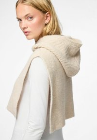 Sudadera sin mangas de punto beige con una tela texturizada, que presenta una capucha acanalada y lados abiertos, llevada sobre una camiseta blanca de manga larga.
