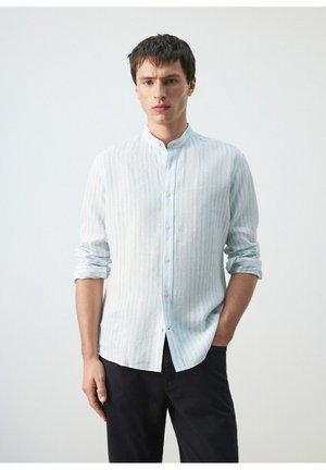 Mężczyzna ubrany w jasnoniebieską i białą, paskowaną koszulę na guziki z podwiniętymi rękawami oraz czarne spodnie, stojący na jednolitym tle.