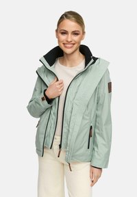 Lachende vrouw in een lichtgroene zip-up jas over een crèmekleurige trui en crèmekleurige broek, met één hand de kraag van de jas vasthoudend.