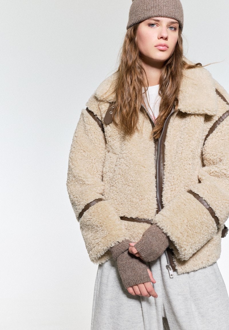 Beige donzige shearling jas met bruine leren accenten, voorzien van een ritssluiting en een brede kraag. Gecombineerd met bruine vingerloze handschoenen.