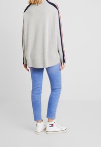 Szary sweter z granatowymi i czerwonymi paskami, w zestawieniu z jasnoniebieskimi obcisłymi dżinsami i białymi sneakersami na wysokiej podeszwie z logo.