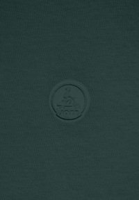 T-shirt vert foncé avec un logo rond embossé au centre. Le tissu semble doux, avec une texture lisse et un design minimaliste.