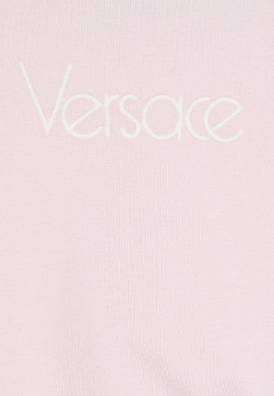 Balta izšūta "Versace" logo uz gaiši rozā auduma fona, kas demonstrē materiāla detalizēto tekstūru.