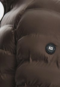 Ciemnobrązowa puchowa kurtka o pikowanej fakturze i widocznych szwach. Posiada okrągłą czarną naszywkę z białym napisem.