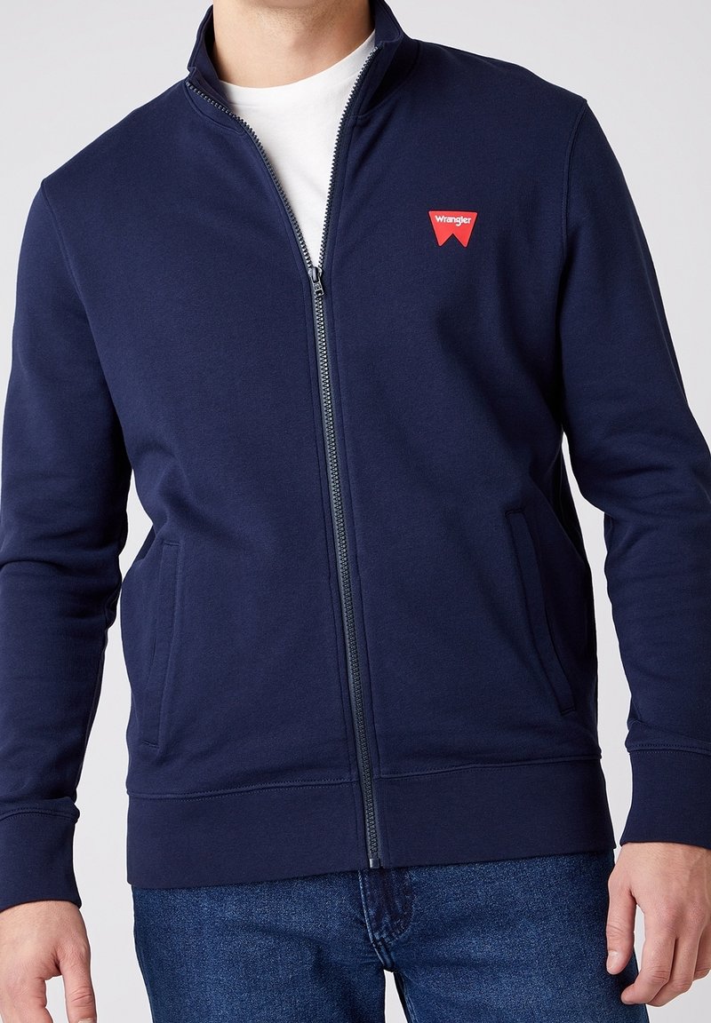 Marineblå zip-jacka i bomullsblandning, med sidfickor och ribbade muddärmar. Wrangler-logotypen visas på övre vänstra bröstet.