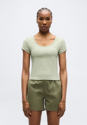 Kvinde med flettet hår iført en lysegrøn tætsiddende T-shirt med korte ærmer og løse olivengrønne shorts, stående mod en ensfarvet baggrund.