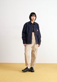 Chaqueta de denim en azul oscuro con un bolsillo en el pecho, combinada con pantalones holgados beiges y zapatillas deportivas negras. Fondo simple.