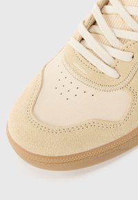 Sneaker bege com uma combinação de camurça e couro liso, com perfurações para ventilação, uma sola texturizada e atacadores creme.