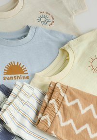 Trois ensembles bébé pliés avec des imprimés à thème soleil et des pantalons à taille élastique dans des motifs beige zigzag et rayures blanches.