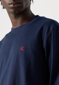Muškarac u tamnoplavom dukserici s malim crvenim vezom 'cK' logotipom na prsima, na jednostavnoj bijeloj pozadini.