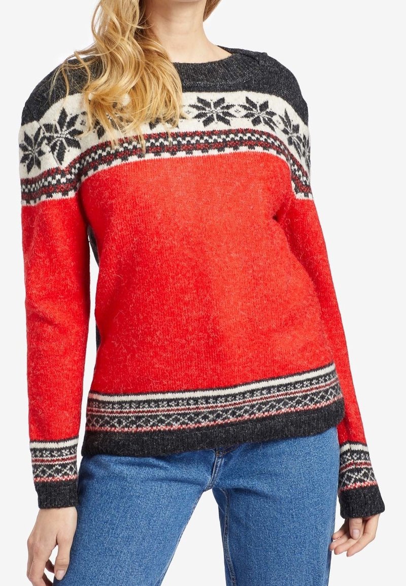 Femme portant un pull en maille rouge, noir et blanc avec un motif flocon de neige, associé à un jean bleu, raccourci au niveau du cou et des cuisses.
