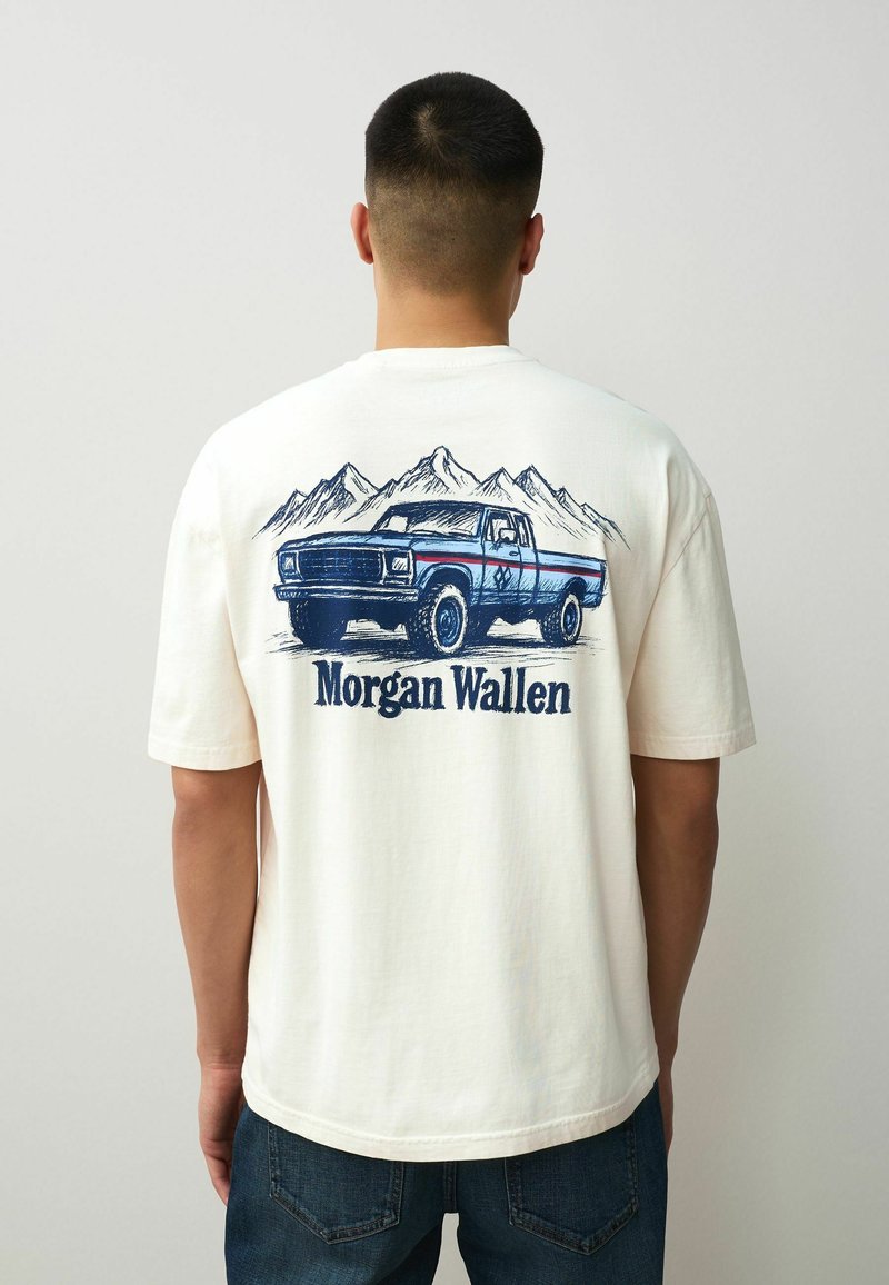 Uomo che indossa una camicia bianca con un design di un pickup vintage blu e montagne, con il testo "Morgan Wallen" sul retro.