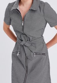 Robe à manches courtes avec un motif géométrique en noir et blanc, dotée d'une fermeture éclair frontale, de poches latérales et d'un détail noué à la taille.