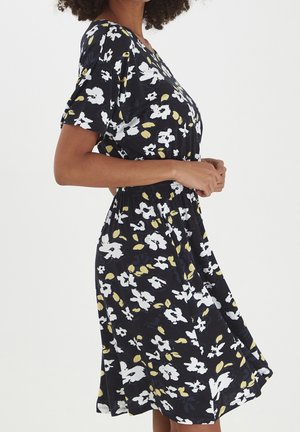 Mujer con un vestido negro con estampados florales blancos y amarillos, con las manos juntas a la altura de la cintura, sobre un fondo claro y liso.