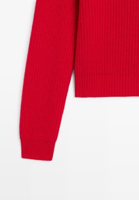 Röd stickad tröja med ribbade ärmslut och texturerat mönster, uppvisad mot en vit bakgrund.
