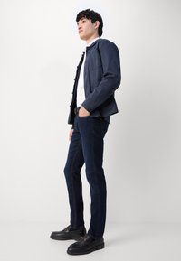 Mornarska jakna preko bijele košulje, uparenih s tamnoplavim trapericama. Outfit uključuje crne cipele s detaljem zlatne kopče. Slim fit silueta.