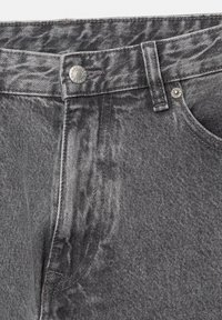 Gros plan sur un jean denim gris montrant la fermeture par bouton, la passants de ceinture, le rivet métallique et les détails de couture de la poche avant.