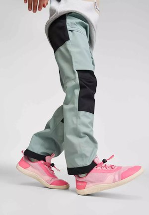 Personne portant un pantalon cargo vert clair et noir avec des chaussures de sport rose vif, marchant sur un fond clair uni.