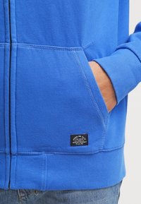 Sudadera azul hecha de una mezcla de algodón con un bolsillo frontal. Presenta una etiqueta negra con texto blanco. La tela muestra una textura suave.