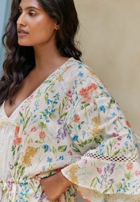 Šviesiai kreminės spalvos kimono su gėlių raštu, kuriame vaizduojamos įvairiaspalvės gėlės, tekstūruotas audinys ir nėrinių apdaila ant rankovių.