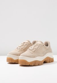 Beige Wildleder-Sneaker mit dicker weißer Zwischensohle und strukturiertem Gummiaußensohle. Verfügt über runde, flache Schnürsenkel und gestickte Details am Obermaterial.