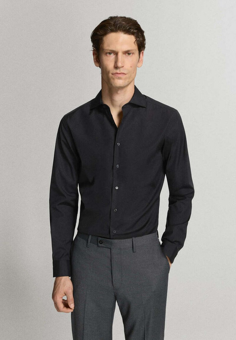 Homme debout avec une main dans la poche portant une chemise noire à manches longues boutonnée et un pantalon gris sur un fond gris clair.