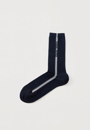 Une chaussette bleu marine à tige mi-haute, avec deux bandes blanches qui partent du bord-côte et longent le pied, ainsi qu’un lettrage vertical sur la tige.