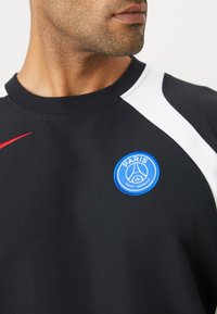 Sudadera negra con cuello redondo, que presenta acentos en blanco en los hombros y un logo circular azul que dice "Paris Saint-Germain" en el pecho.