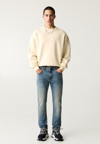 Crèmekleurig sweatshirt, lichtblauwe jeans en donkere sneakers. Het sweatshirt heeft een relaxte pasvorm; de jeans vertonen lichte vervaging; de sneakers hebben meerkleurige accenten.