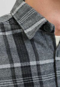 Camisa de cuadros en gris y negro con una tela texturizada, que presenta un collar con botones y franjas blancas en contraste en el patrón.
