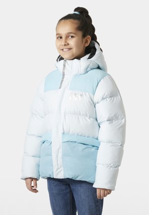 Jong meisje dat lacht, gekleed in een lichtblauwe en witte gewatteerde winterjas met kap en een blauwe spijkerbroek, staand tegen een witte achtergrond.