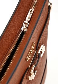 Borsa a tracolla in pelle marrone con texture, dotata di hardware dorato, chiusura con zip e logo del marchio visibile in rilievo sul lato.