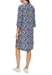 Kleid mit Blumenmuster in Blau-, Rosa- und Schwarztönen. Mit Dreiviertelärmeln, seitlichen Schlitzen und lockerem Schnitt. Getragen mit weißen Sneakers.