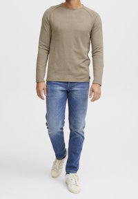 Chemise à manches longues couleur marron clair avec un col rond et des poignets côtelés, portée avec un jean bleu délavé et des baskets blanches.