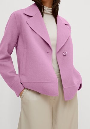 Kvinde iført en lyserød uldjakke over en hvid rullekrave, med hænderne i lommerne på beige bukser med brede ben.