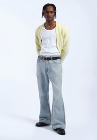 Puszysty jasnożółty kardigan na białej bluzce bez rękawów, w połączeniu z jasnoniebieskimi dżinsami w stylu dzwonów i czarnymi butami. Minimalne widoczne akcesoria.