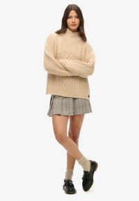 Beige gebreide trui met hoge kraag en kabelpatroon, gecombineerd met een geruite plooienrok, kniehoge sokken en zwarte schoenen.