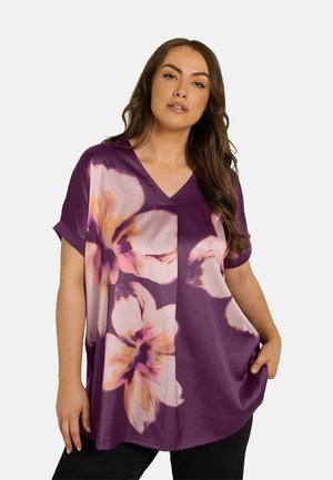 Frau mit langen braunen Haaren, die ein lila V-Ausschnitt-Top mit großen rosa und orangen Blumenmustern trägt und den Saum mit einer Hand hält.