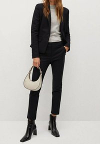 Blazer nero sopra un maglione grigio, abbinato a pantaloni neri su misura. Tiene una piccola borsa color crema e indossa stivaletti neri alla caviglia.