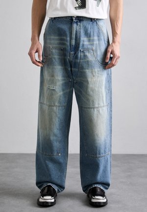 Wide-leg blue denim jeans with a faded finish, distressed patches, large pockets, and visible stitching details; styled with black shoes.