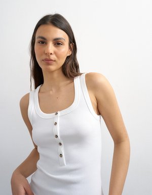 Jeune femme aux longs cheveux foncés portant un haut blanc sans manches en maille côtelée avec quatre boutons, debout devant un fond blanc uni.