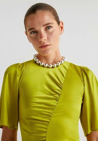 Abito aderente verde brillante con una cucitura laterale teksturata, maniche a sbuffo corte e una collana di perline argento lucido attorno al collo.