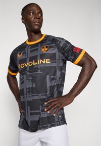 Mand i sort og gråt mønstret sportsjakke med orange detaljer og "NOVOLINE" logo, poserende med hænderne på hofterne, mens han ser til siden.