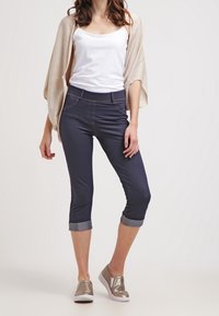 Mörkblå cropped byxor med uppvikta fållar, featuring fem fickor och guldstickning, i kombination med en vit linne och en beige cardigan.