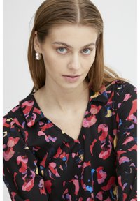 Schwarzes Shirt mit buntem abstraktem Blumendesign, mit V-Ausschnitt und langen Ärmeln. Das Model trägt silberne ovale Ohrringe und hat ein minimales Make-up.