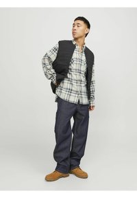 Schwarze gesteppte Weste über einem karrierten Hemd in Blau-, Grün- und Weißtönen, kombiniert mit lockeren dunklen Jeans und braunen Schuhen.