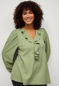 Blouse verte texturée avec un décolleté en V, présentant un devant volants et des manches longues. Conçue avec une coupe ample et des détails subtils.